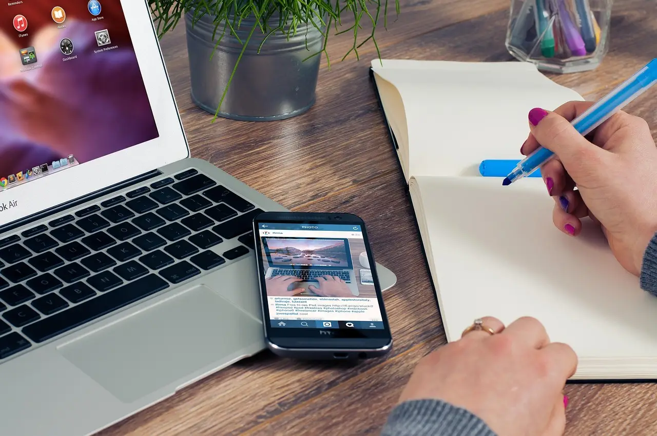 A laptop, a phone showing a photo app, and a hand writing in a notebook on a wooden desk