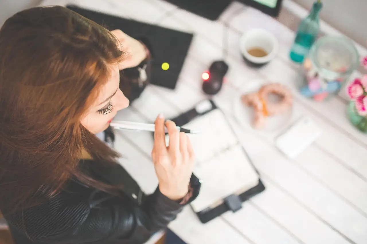 Overhead view of a woman writing in a planner with a pen beside coffee on a white table