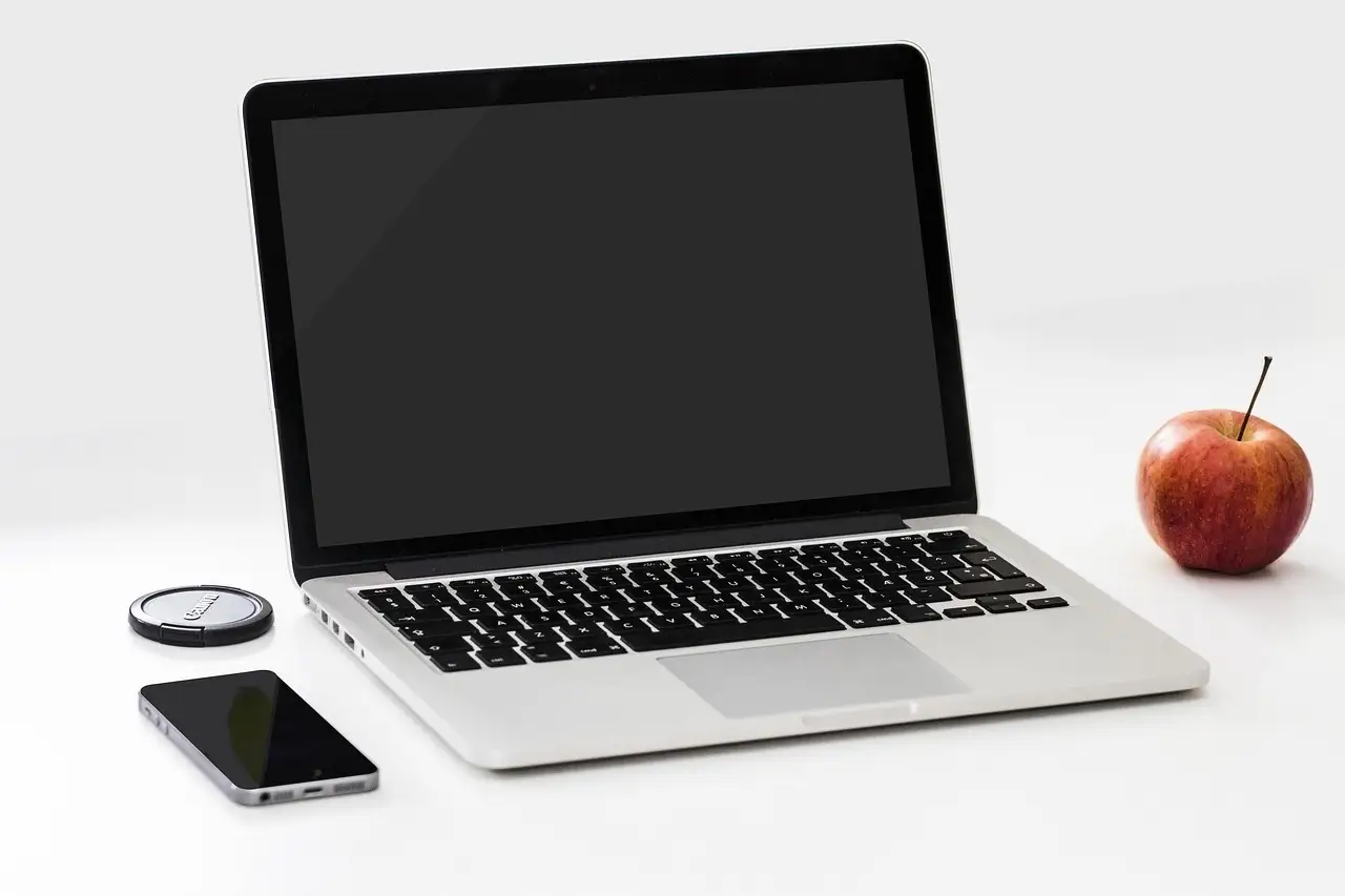 An open laptop with a blank screen beside a smartphone, a camera lens cap, and a red apple on a white surface