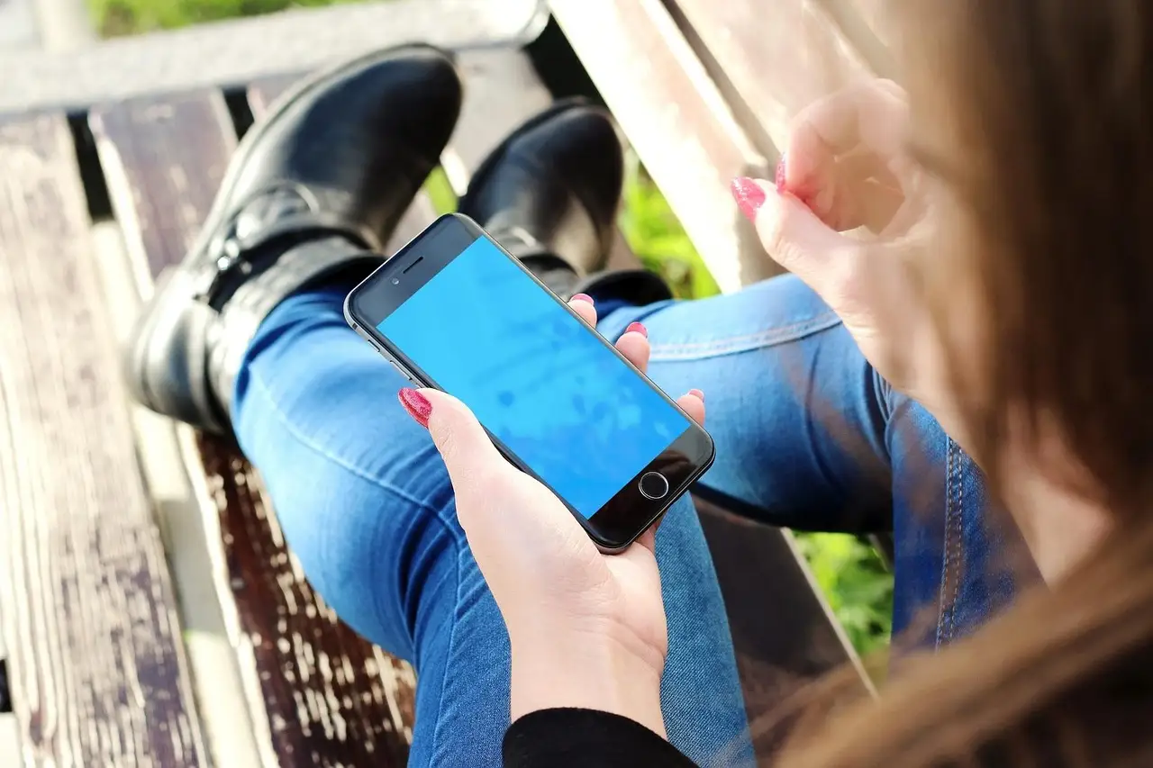 A woman sitting on a bench outdoors holds a black smartphone with a blue screen