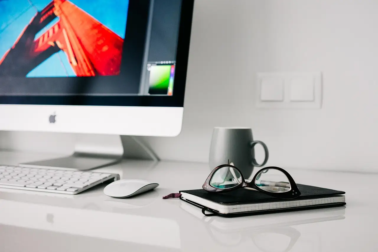 An iMac, wireless keyboard and mouse, and glasses on a notebook beside a mug on a white desk